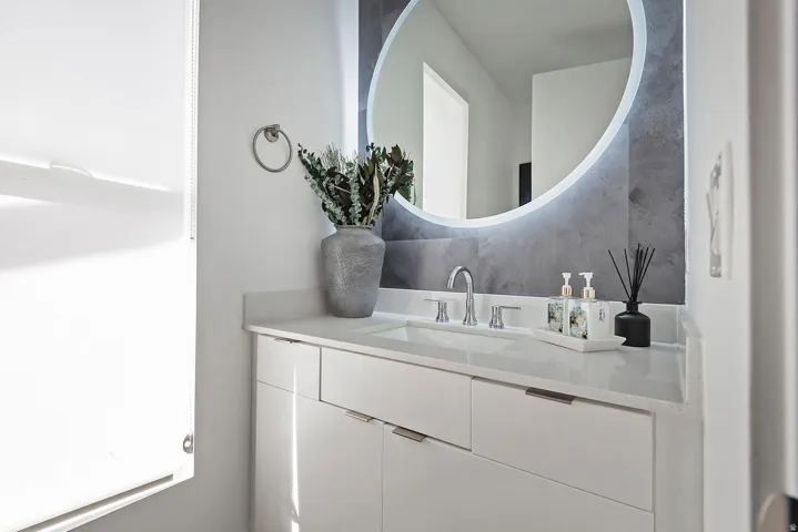 White vanity with integrated sink, modern chrome faucet, light-toned countertop, and an illuminated circular mirror