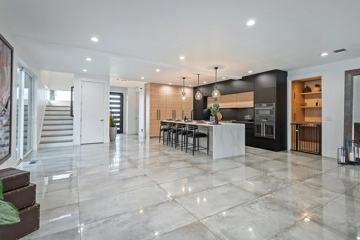 Spacious open-concept interior with large format floor tiles, a contemporary kitchen featuring integrated appliances, a central island with bar seating, and a sleek dark entry door