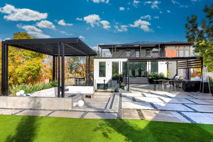 Expansive artificial turf lawn, large concrete paver patio with dark stone infill, multiple contemporary pergolas, built-in concrete planters, and a multi-level architectural design