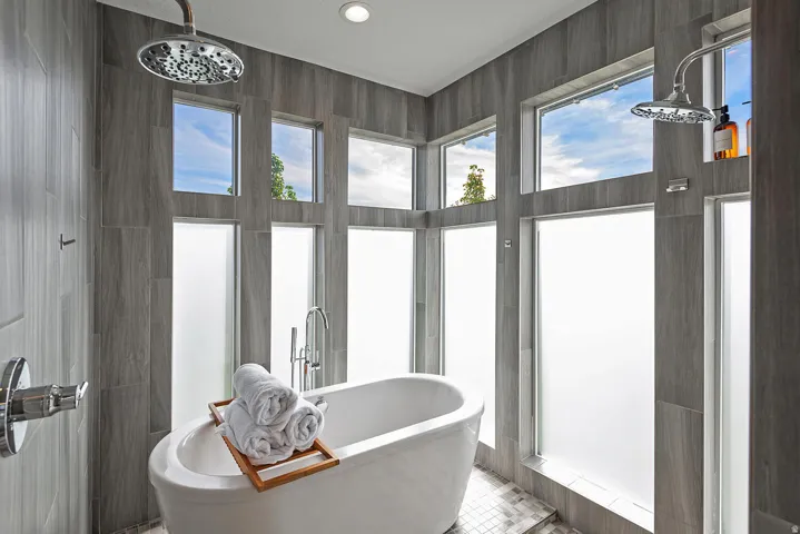 Freestanding soaking tub with chrome bath filler, surrounded by large format tile, dual rain shower heads, and multiple windows