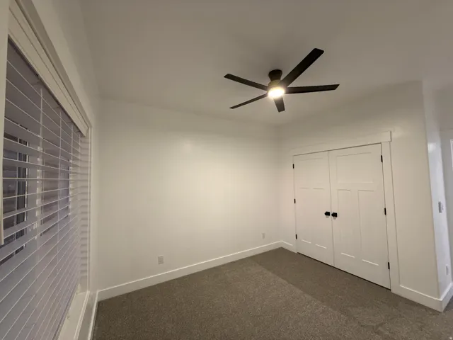 White walls, carpeted flooring, window with horizontal blinds, ceiling fan with integrated lighting, and white bi-fold closet doors with black hardware