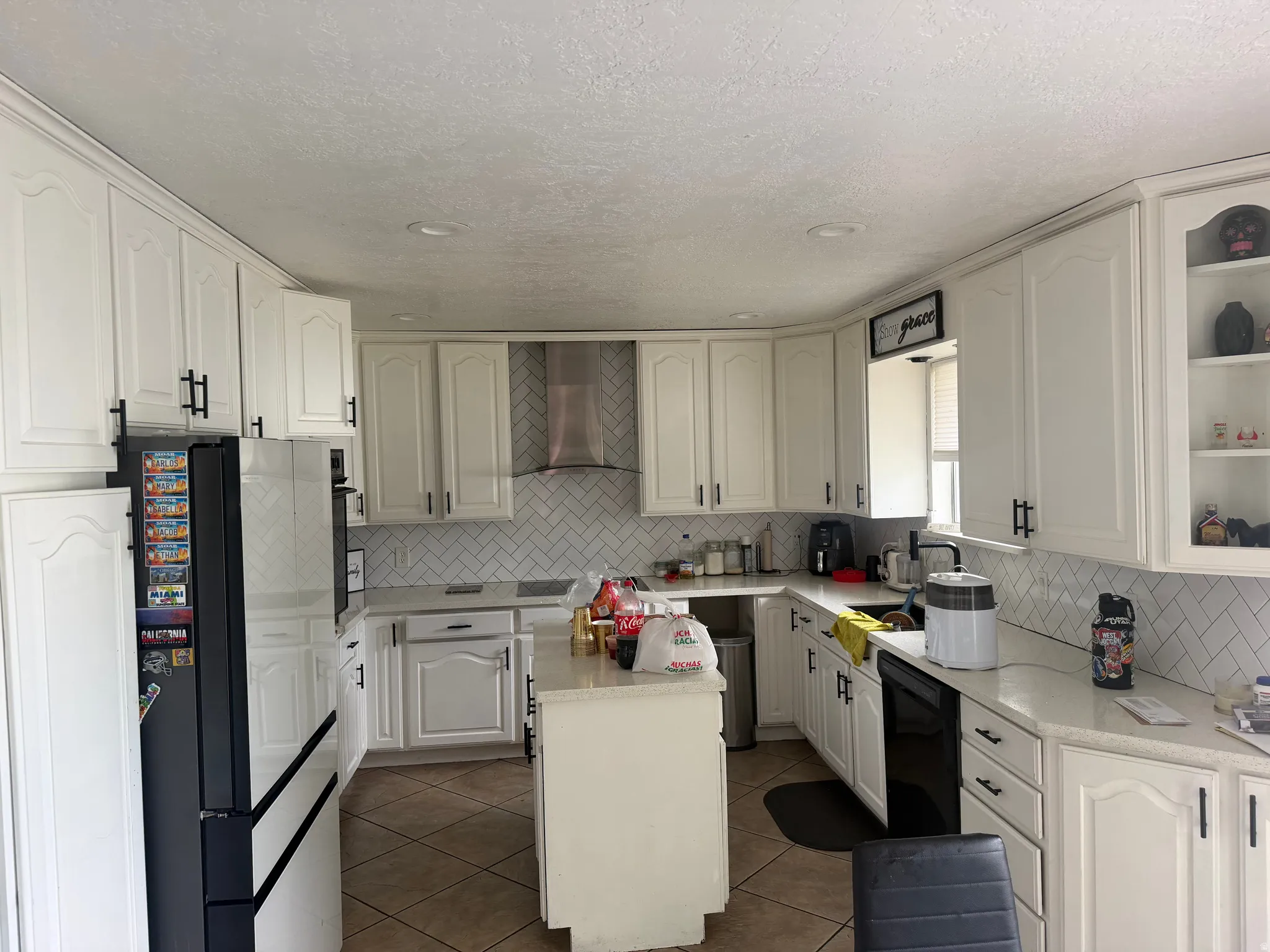 White cabinetry with black hardware, white herringbone tile backsplash, integrated stainless steel hood, recessed lighting, and tile flooring