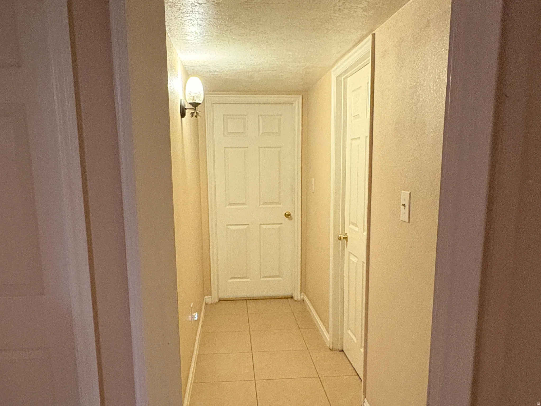 Hallway featuring tile flooring, neutral wall tones, white paneled doors, and wall-mounted lighting