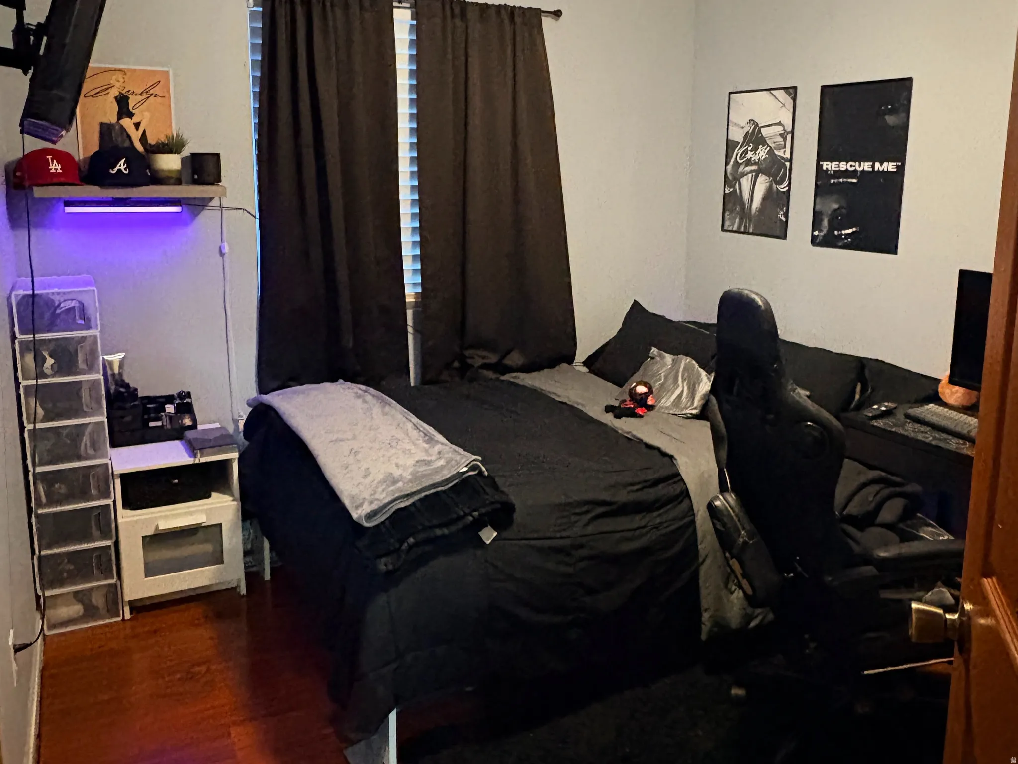 Wood-finish flooring, ceiling fan with integrated lighting, window with blinds, wall-mounted shelving, and a wall-mounted television bracket