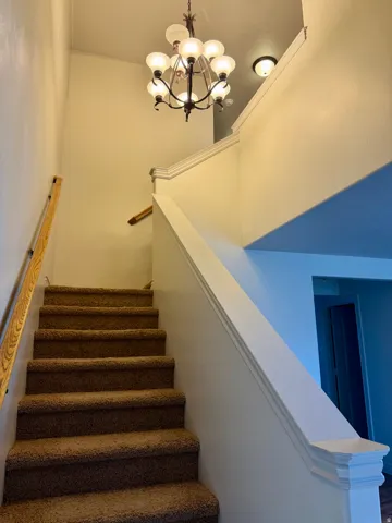 Carpeted staircase with a wood handrail, white painted walls, and a multi-bulb chandelier