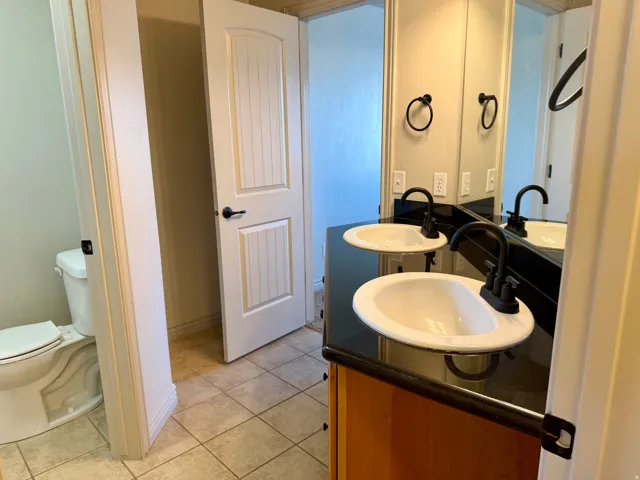 Dual white oval sinks, black countertop, matte black fixtures, wood-finish vanity, and tiled flooring