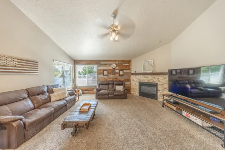 Vaulted ceiling with a ceiling fan, stone fireplace with a wood mantel, wood-finish accent wall, and multiple windows
