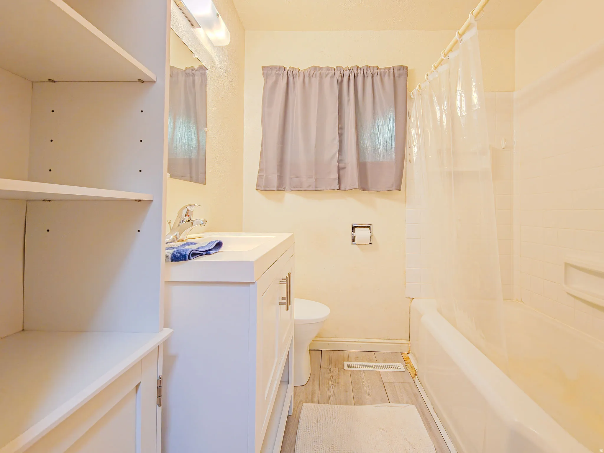 Bathroom featuring a white vanity with integrated sink, chrome faucet, white tub with tiled surround, wood-finish flooring, and built-in shelving