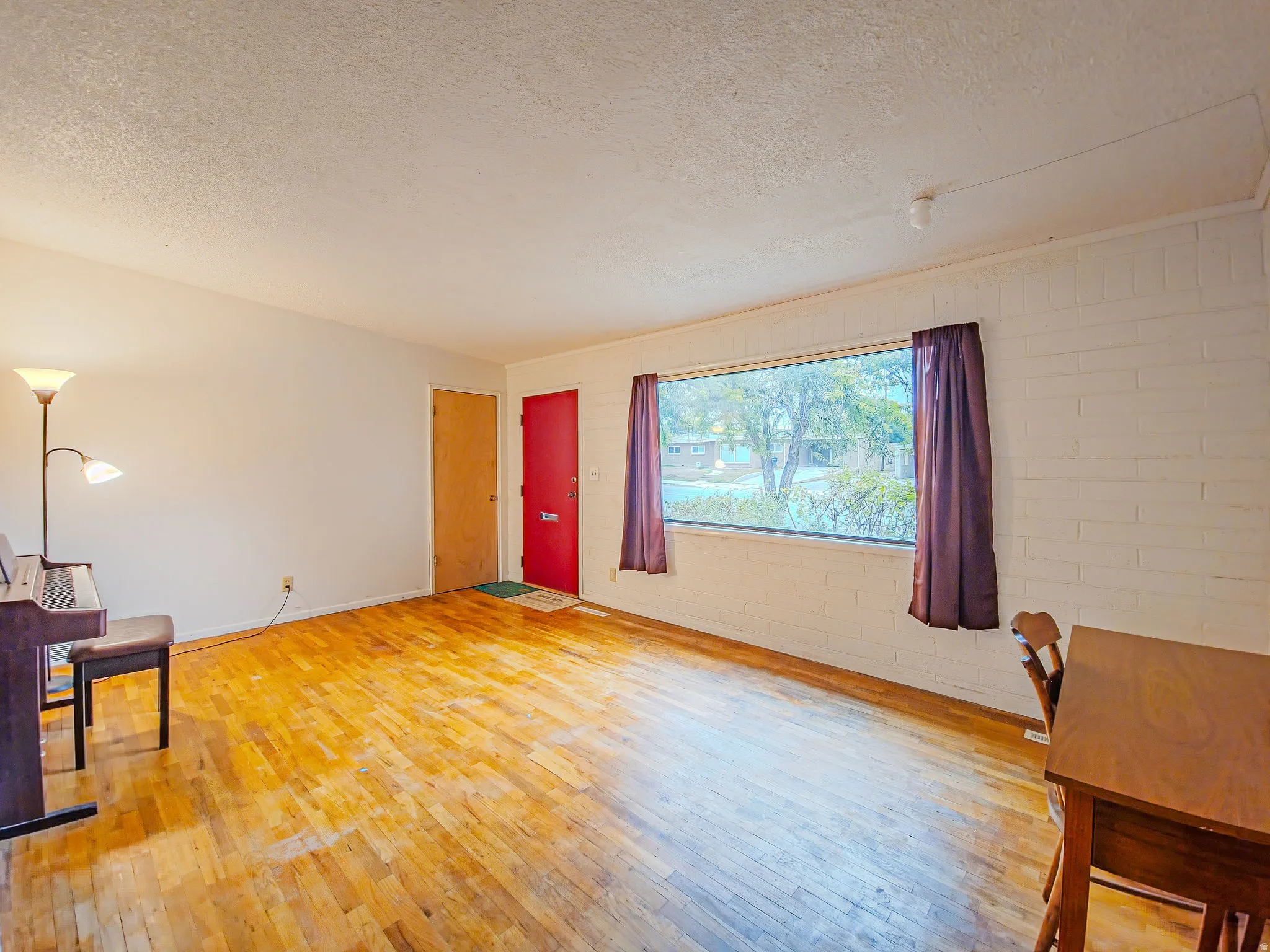 Hardwood flooring, painted brick accent wall, large window, red entry door, and a flush-mounted ceiling light