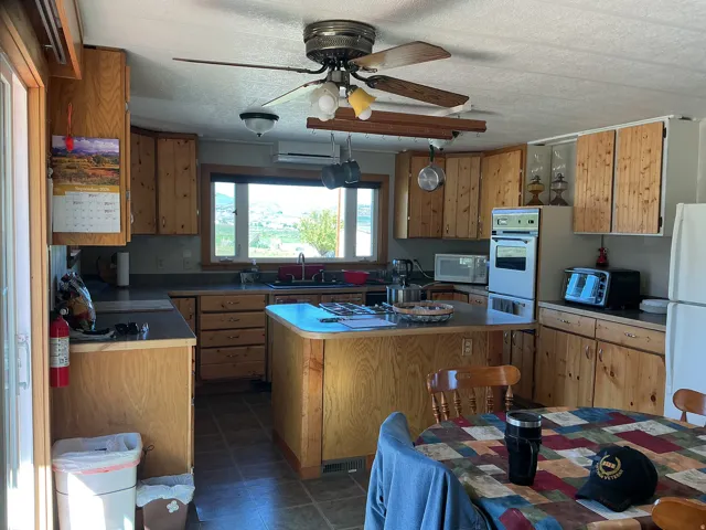 Wood cabinetry with dark countertops, built-in double oven, ceiling fan with integrated lighting, and a kitchen island