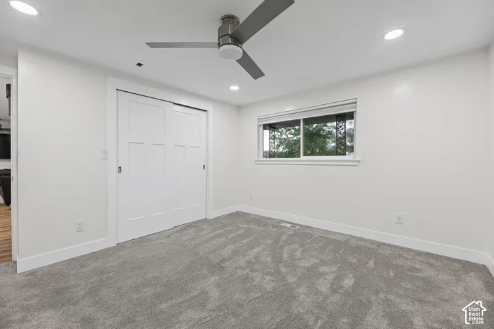 Recessed ceiling lighting, gray carpeting, a window with a roller shade, a ceiling fan, and white sliding closet doors