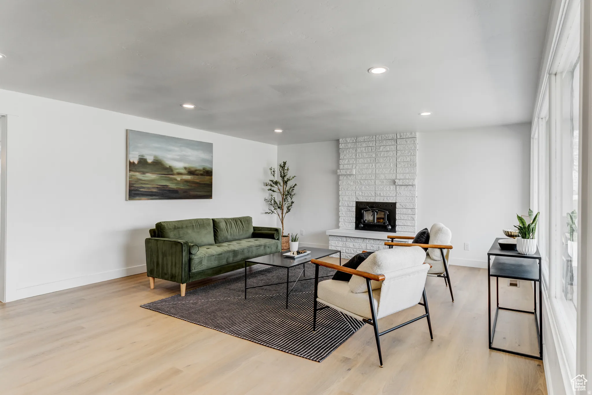 Light wood-finish flooring, white walls, recessed ceiling lighting, a brick fireplace, and tall windows
