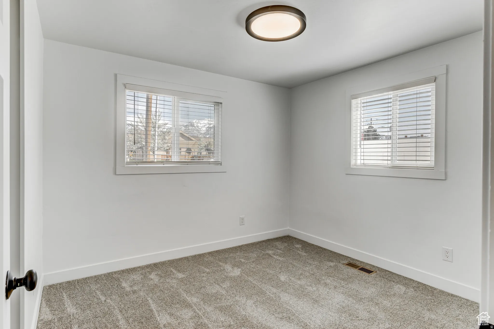 Neutral wall finishes with a ceiling-mounted light fixture, two windows featuring white blinds, and gray patterned carpet