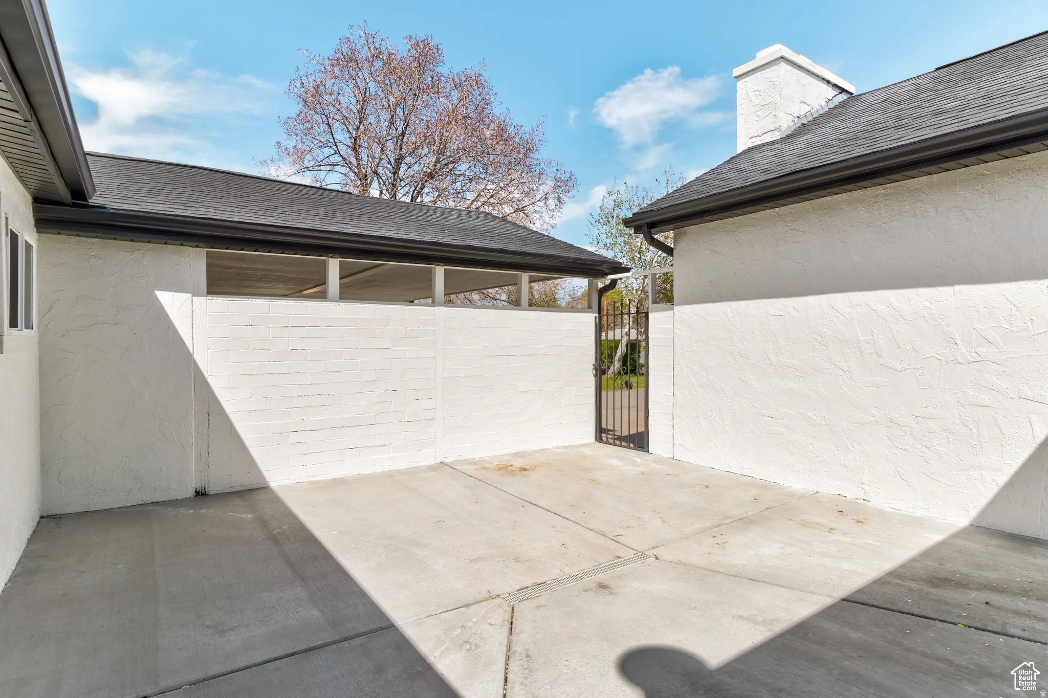 Private paved courtyard with a wrought iron gate, textured white stucco walls, and a black shingle roof