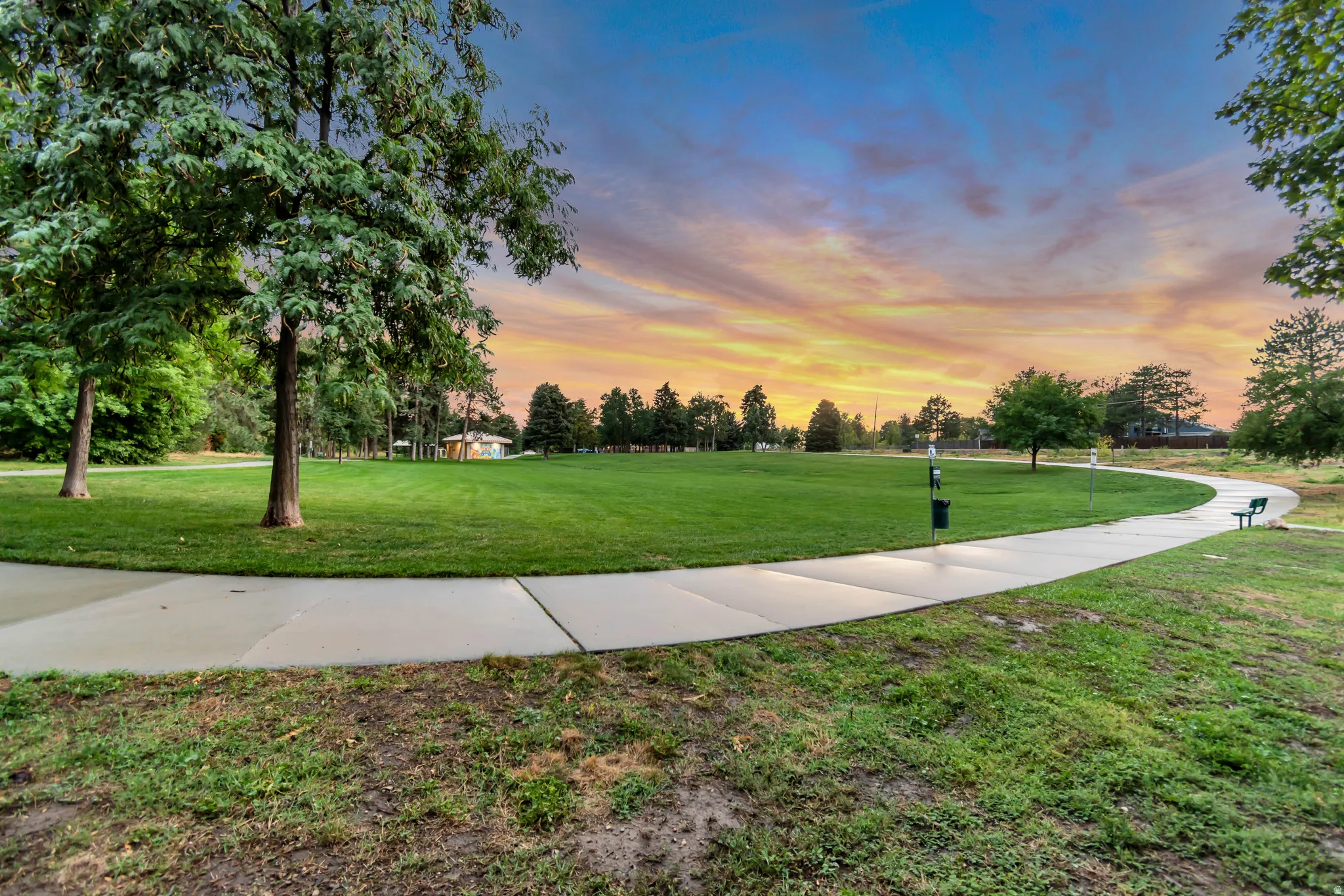 Expansive green space featuring a paved walking path, mature trees, and a distant building structure