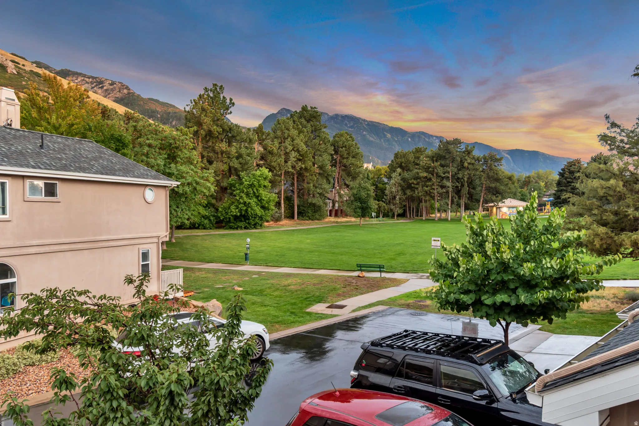 Paved driveway with adjacent green space featuring a bench and paved pathways, surrounded by mature trees and mountain scenery