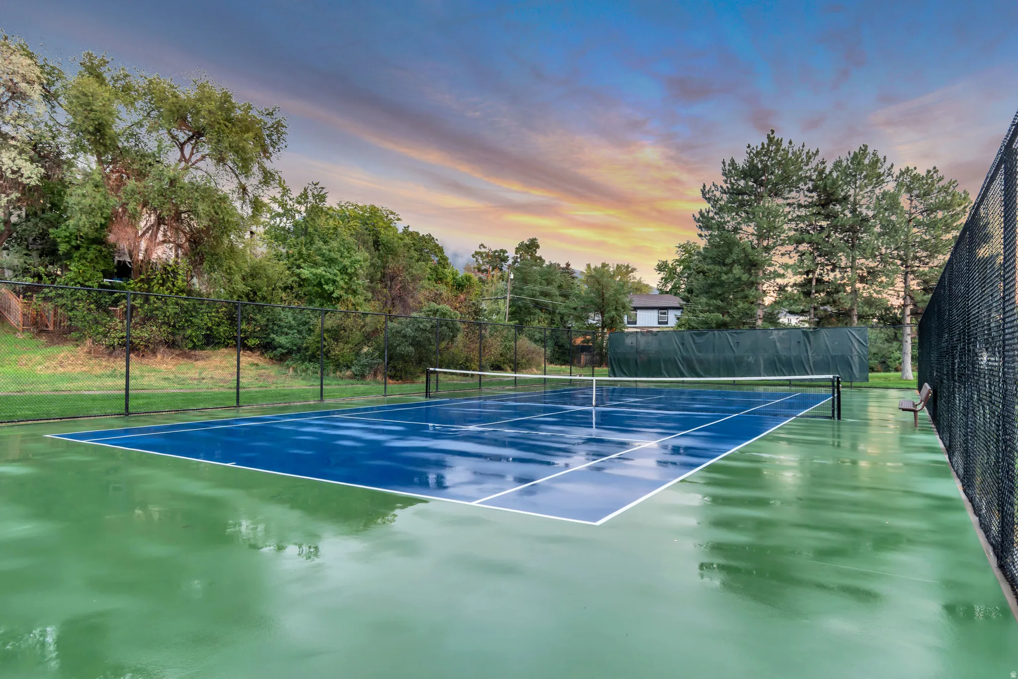 Private fenced tennis court featuring a blue playing surface, green perimeter, and surrounding mature trees
