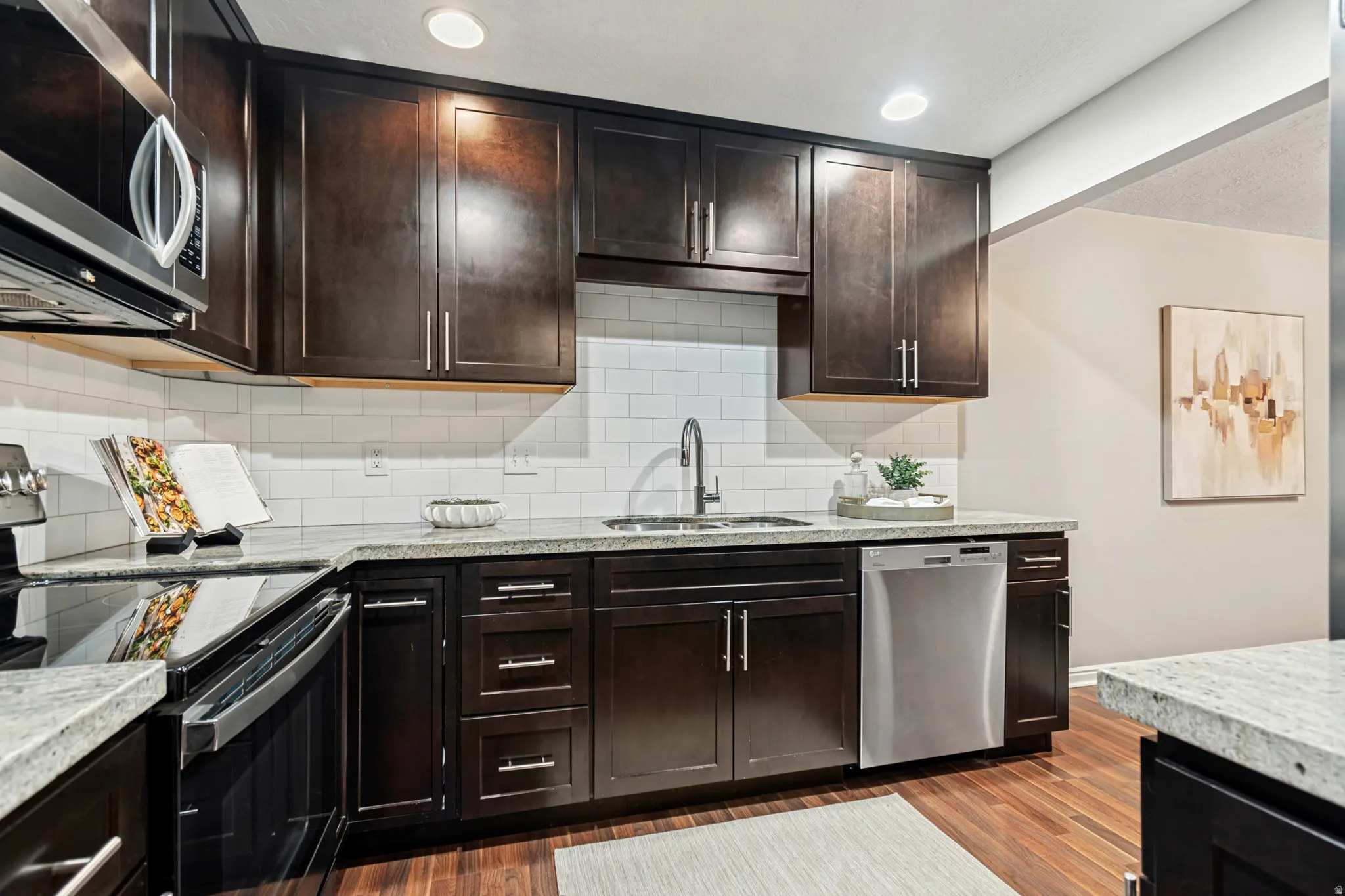 Kitchen featuring dark wood cabinetry, light-toned countertops, a white subway tile backsplash, stainless steel appliances, and wood-finish flooring