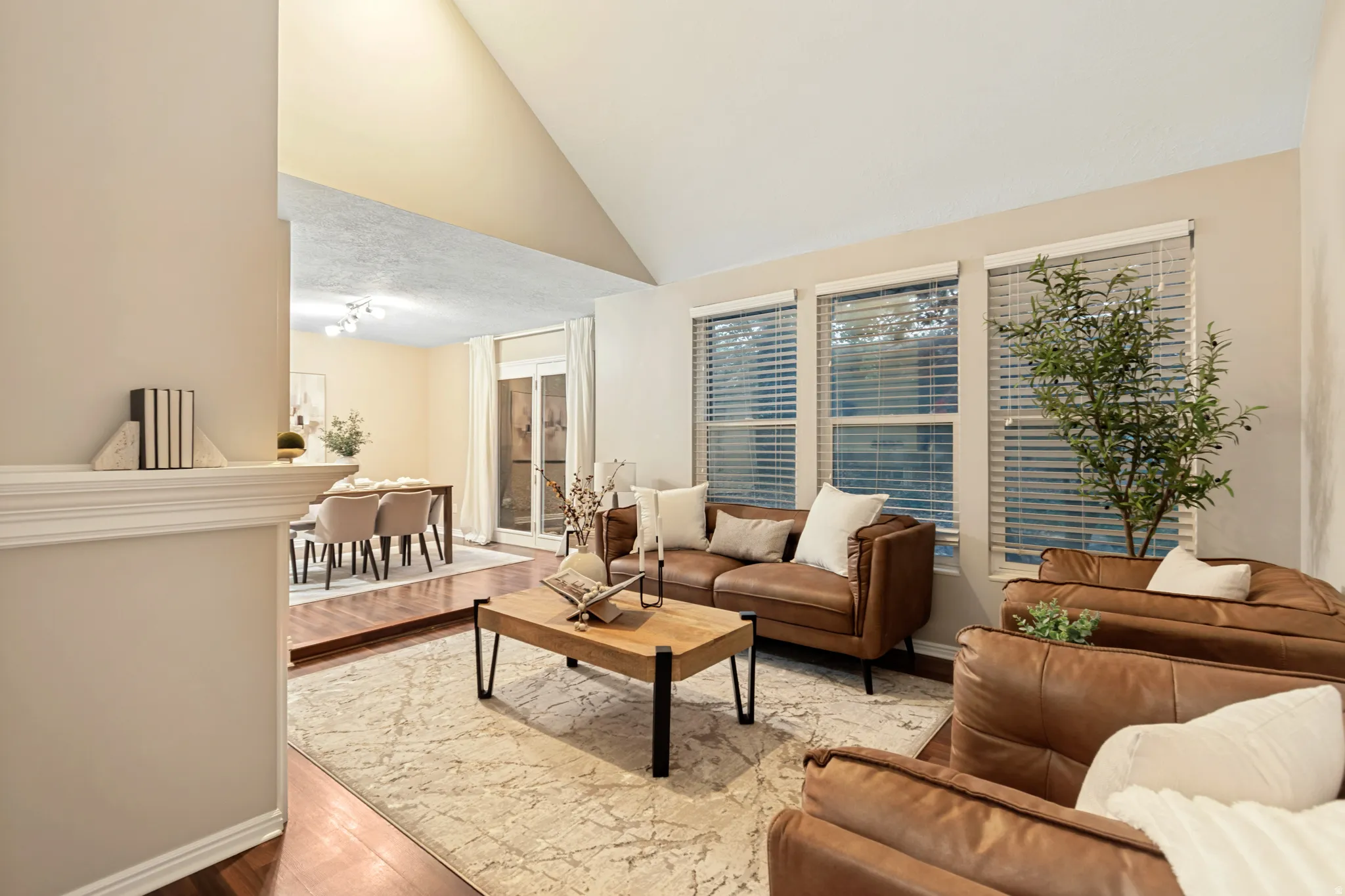 Vaulted ceiling, multiple windows with blinds, wood-finish flooring, and an open concept layout with an adjacent dining area featuring a sliding glass door