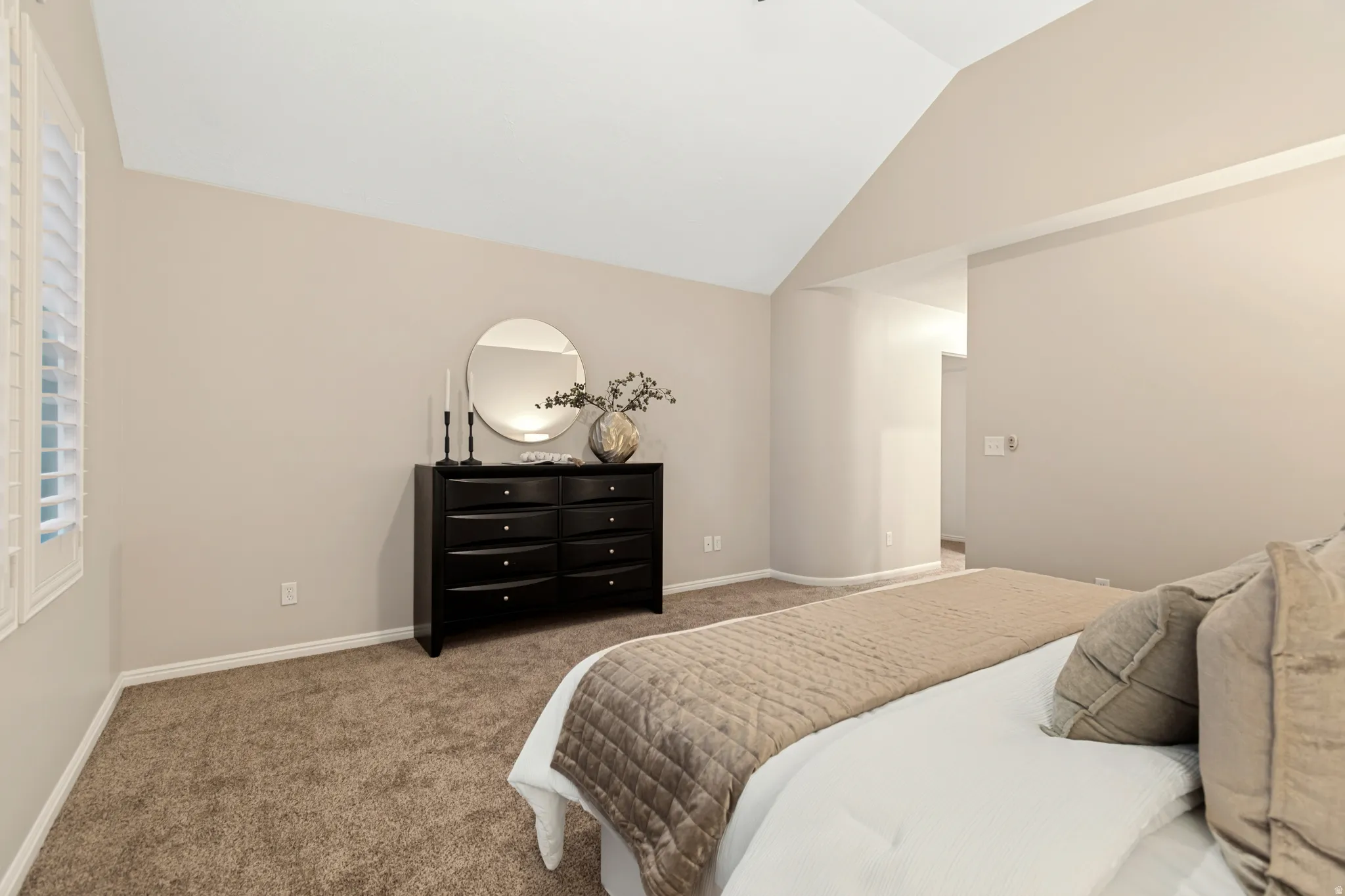 Vaulted ceiling with neutral wall tones, carpeted flooring, and window with plantation shutters