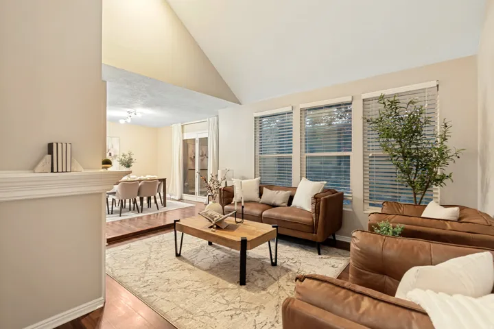 Vaulted ceiling, multiple windows with blinds, wood-finish flooring, and an open concept layout with an adjacent dining area featuring a sliding glass door