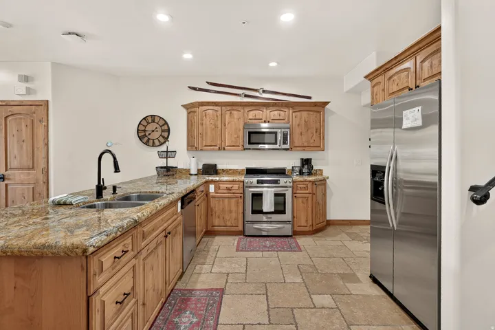 Kitchen featuring extensive wood cabinetry, stone countertops, stainless steel appliances, a double basin sink with a matte black faucet, and stone tile flooring