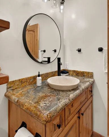 Wood-finish vanity with paneling and black hardware, granite countertop, vessel sink, matte black faucet, and a round black-framed mirror