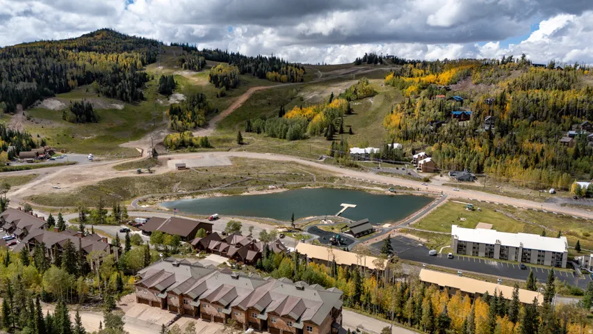 Mountainous terrain with lush coniferous and deciduous trees featuring autumn foliage, a central pond with a dock, and multiple residential structures