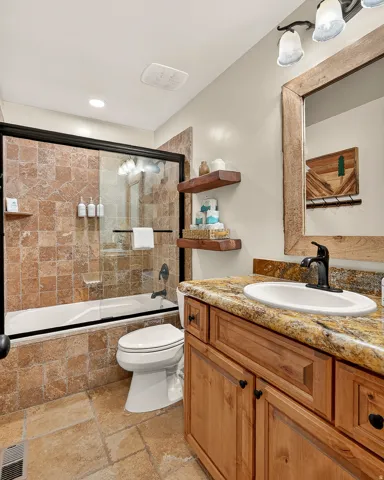 Tiled shower-tub combination with sliding glass doors, natural wood-finish vanity with an undermount sink, stone-finish countertop, and decorative wall mirror