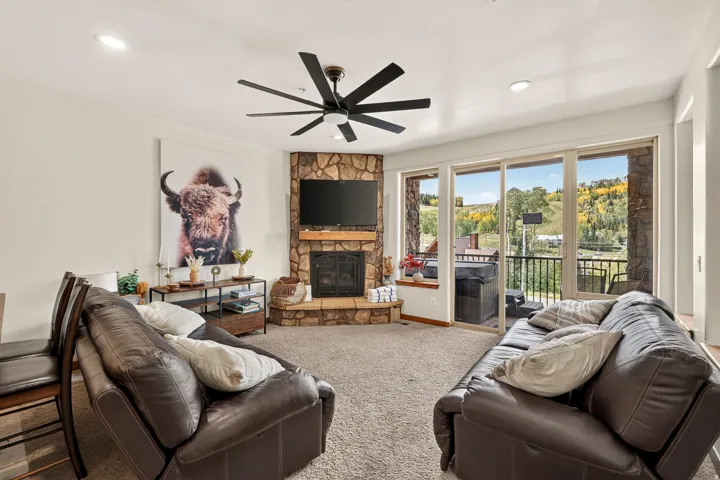 Stone-clad corner fireplace, wall-mounted television, ceiling fan, recessed lighting, and sliding glass doors opening to a balcony