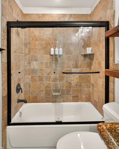 Bathtub with full height tile surround, sliding glass enclosure, oil-rubbed bronze fixtures, and integrated wall shelf