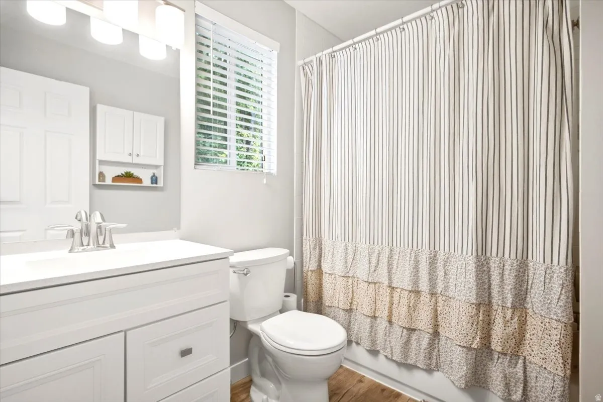 White vanity with integrated sink, chrome faucet, window with blinds, recessed wall cabinet, and wood-finish flooring