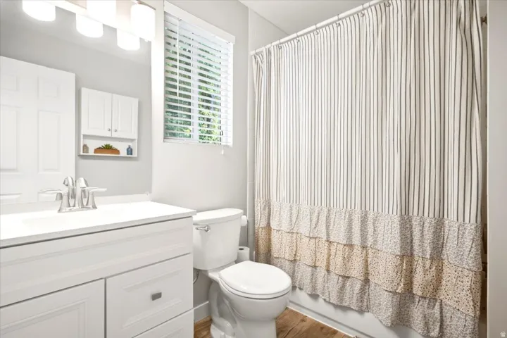 White vanity with integrated sink, chrome faucet, window with blinds, recessed wall cabinet, and wood-finish flooring