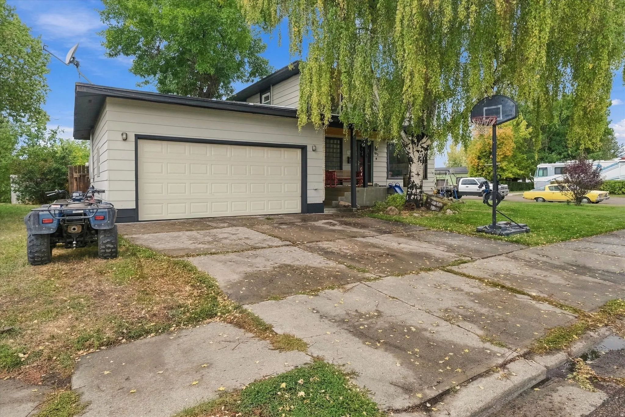 Cream-colored horizontal siding, attached garage with panel door, concrete driveway, front yard with mature trees, and a small front porch