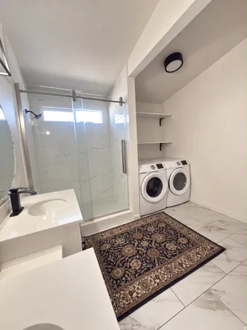 Modern bathroom featuring a dual vanity with white countertops, a walk-in shower with a glass enclosure, white tile flooring, and integrated laundry appliances