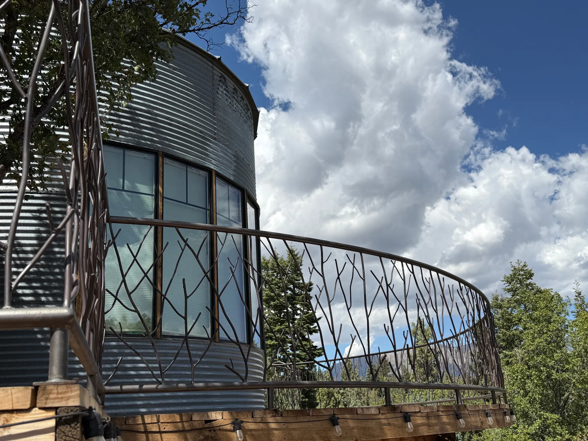 Curved exterior featuring corrugated metal siding, custom branch-patterned metal railing, wood-frame windows, and an outdoor string light fixture