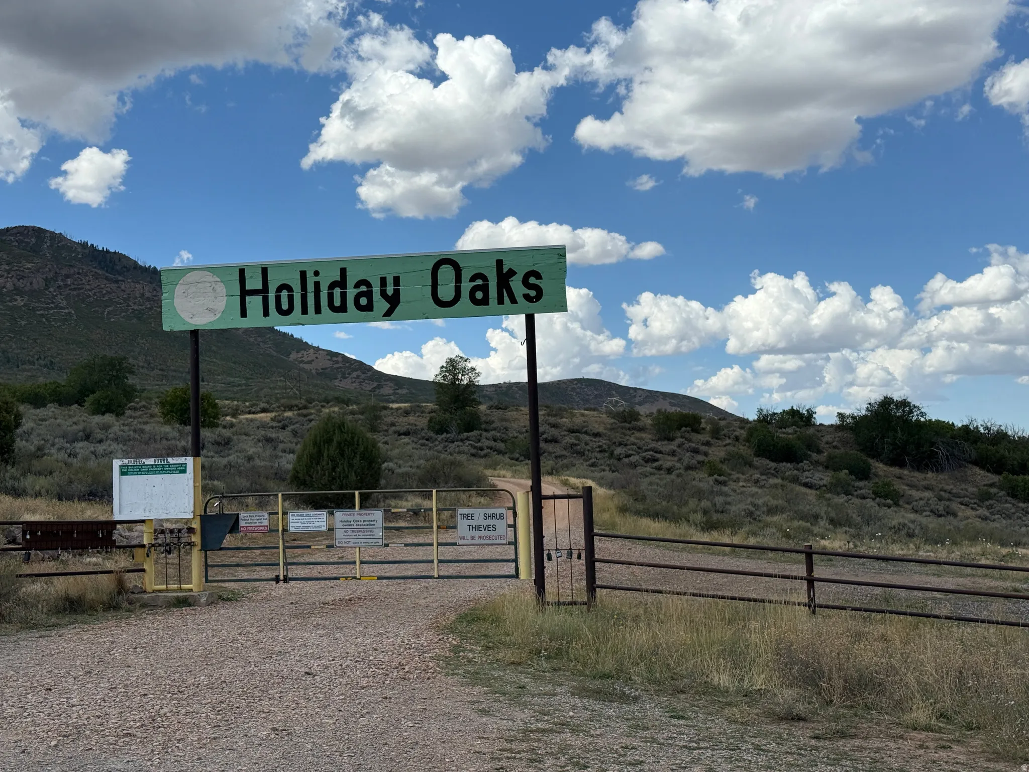 Gated entrance with a "Holiday Oaks" sign, surrounded by natural landscaping and distant mountains