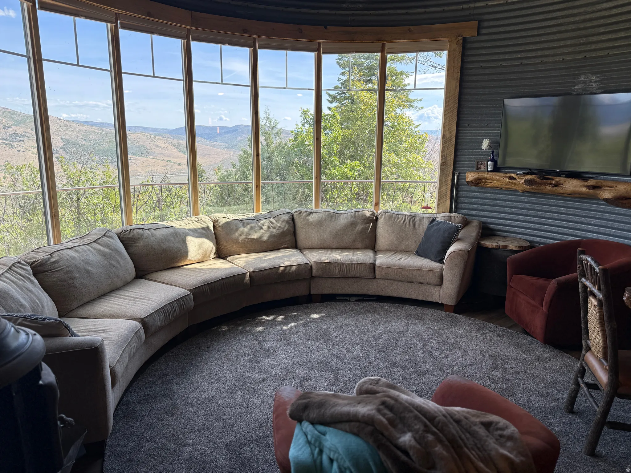 Curved wall of windows, corrugated metal wall paneling, built-in live edge wood shelving, wall-mounted television, and wood-finish window frames