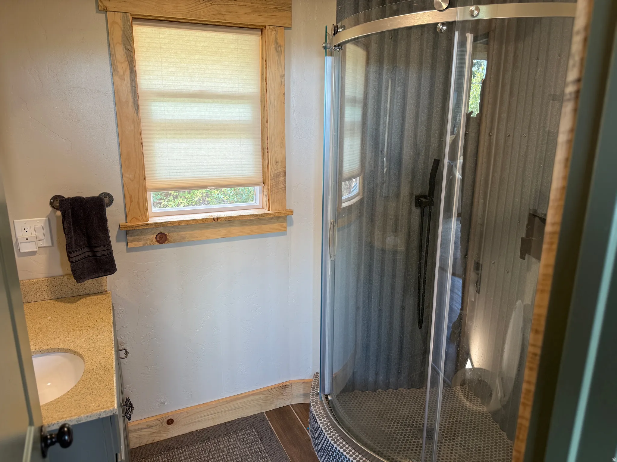 Curved glass shower enclosure with corrugated wall paneling, vanity with an undermount sink and granite-finish countertop, and wood-framed window