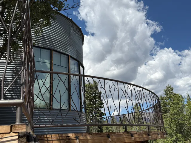 Curved exterior featuring corrugated metal siding, custom branch-patterned metal railing, wood-frame windows, and an outdoor string light fixture