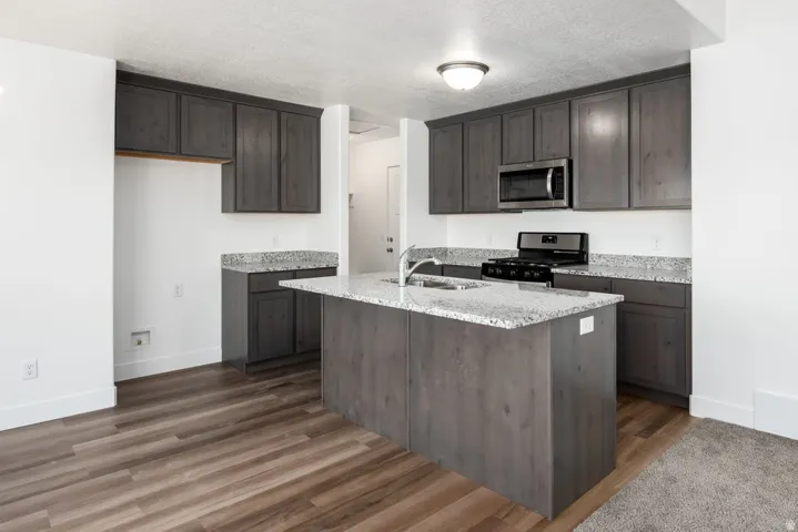 Kitchen featuring wood-finish flooring, dark wood cabinetry, light speckled countertops, a center island with sink, and stainless steel appliances