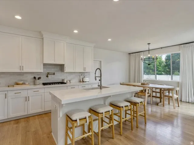 White shaker cabinetry, light-toned wood-finish flooring, recessed lighting, substantial kitchen island with seating, and a gas range with a pot filler