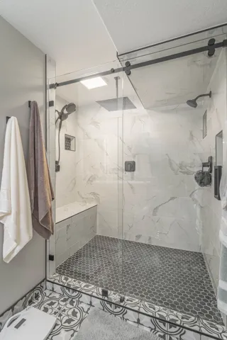 Frameless glass shower enclosure with matte black hardware, white marbled wall tiles, black hexagonal shower floor tiling, built-in shower bench, and a square rain shower head