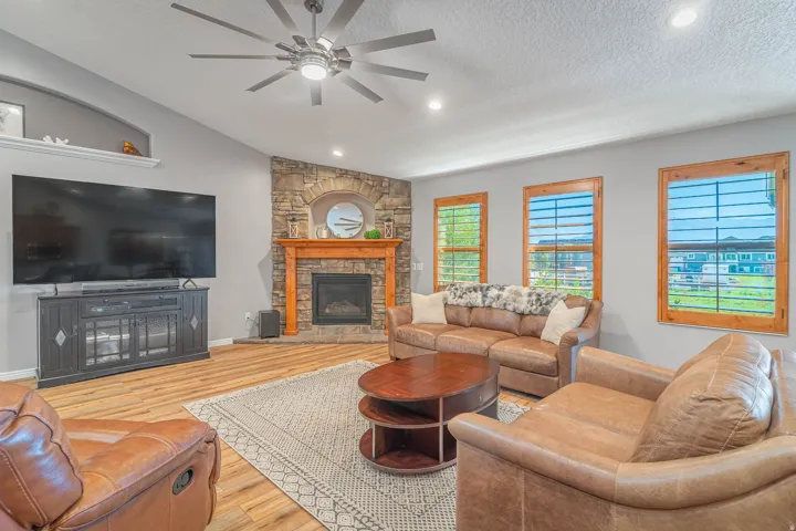 Vaulted ceiling with a contemporary ceiling fan, stone-clad fireplace with a wood mantel, wood-finish flooring, recessed lighting, and multiple windows with wood frames