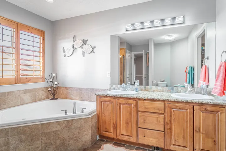 Built-in corner soaking tub with tile surround, wood-finish plantation shutters, double vanity with two undermount sinks, wood-finish cabinetry, and a large wall-mounted mirror