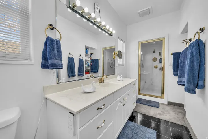 Double vanity with a light-toned countertop, vanity mirror with overhead lighting, built-in shelving, and a glass-enclosed shower with a gold-tone frame