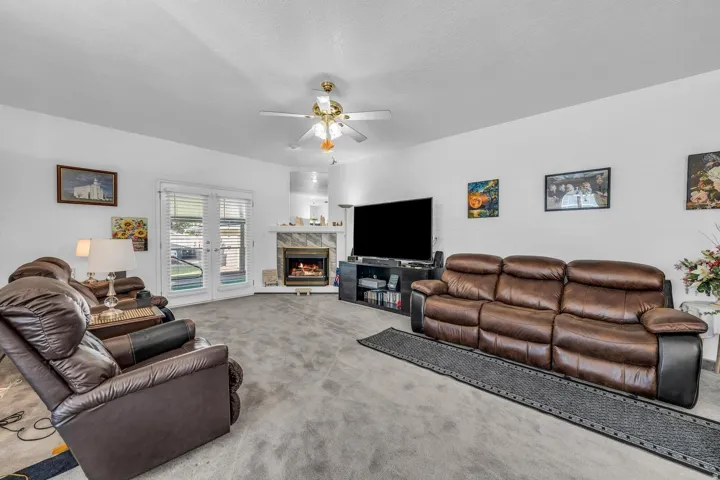 Living area featuring a textured gray carpet, corner fireplace with a tiled surround, white French doors, and a ceiling fan with integrated lighting