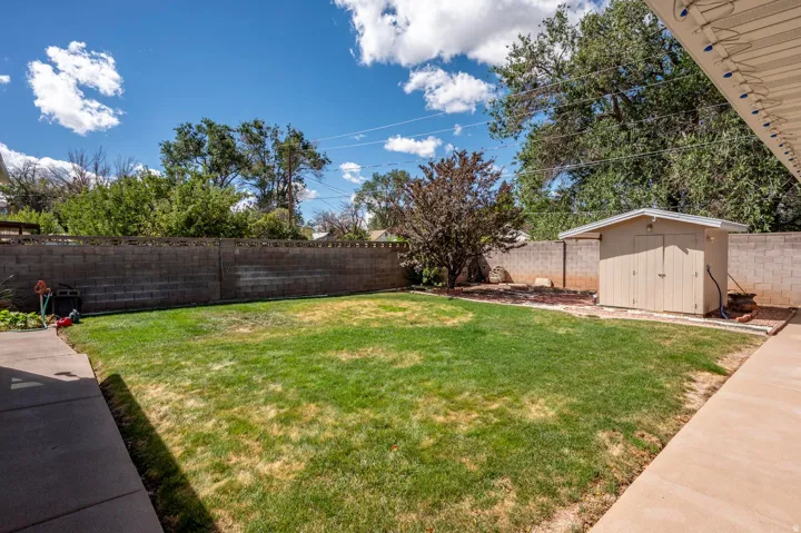 Expansive backyard featuring a manicured lawn, concrete patio, block wall perimeter, mature trees, and a detached storage shed