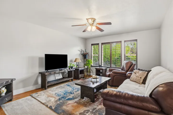Spacious living area featuring wood-finish flooring, multiple windows with plantation shutters, and a ceiling fan with integrated lighting