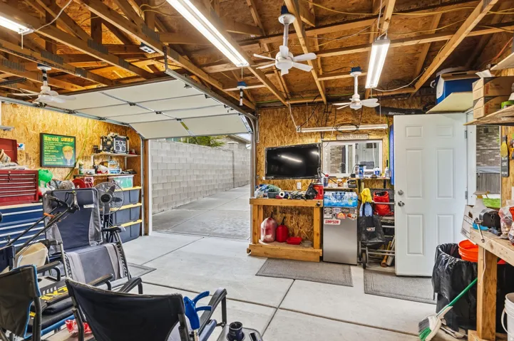 Exposed wood beam ceiling with multiple ceiling fans, overhead fluorescent lighting, concrete flooring, and a roll-up garage door